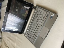 Srebrny laptop HP Mini 210 z lekko podniszczoną klawiaturą stoi na jasnym materiale, ekran odbija otoczenie.