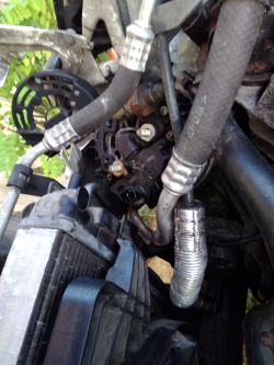 Wymontowany regulator alternatora widoczny w komorze silnika samochodu, otoczony rurkami i przewodami.