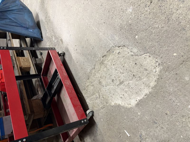 Chropowata betonowa posadzka z jaśniejszą łatą obok czerwonego wózka i desek