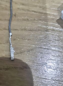 Metalowy pin z przewodem leżący na drewnianej powierzchni, widoczne zaczepy blokujące.
