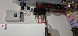 Sterownik Tech podłączony w trybie OpenTherm na ścianie z instalacją hydrauliczną i kolorowymi klapkami.