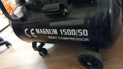 Czarny kompresor powietrza z napisem MAGNUM 1500/50 DENT COMPRESSOR na boku, stojący na podłodze.