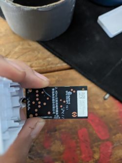 Hand holding a small black PCB with printed markings, solder pads, and a QR code label.