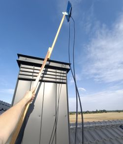 Osoba trzymająca drewnianą tyczkę z anteną na kominie budynku.