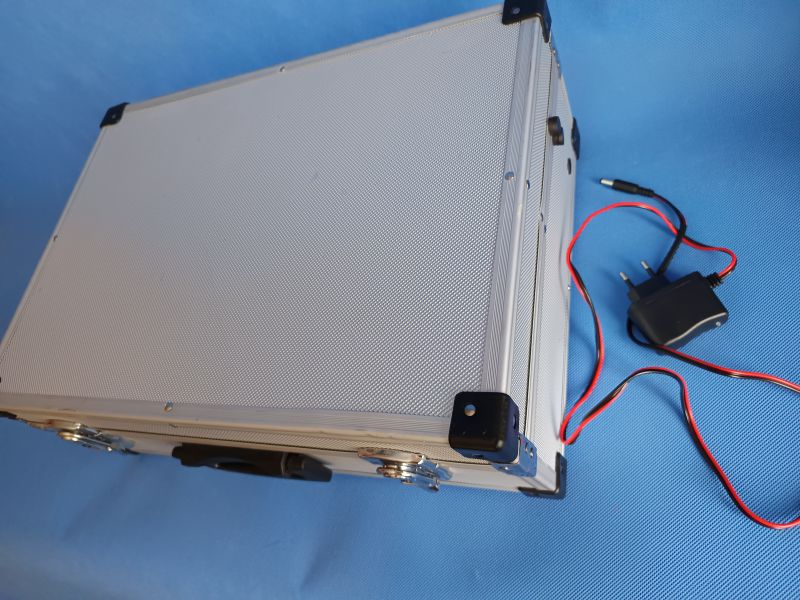 Aluminum case with power adapter Closed silver aluminum case with black corners, next to it a power adapter with wires on a blue background.