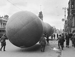 Czarno-białe zdjęcie przedstawiające grupę ludzi ciągnących duży balon ulicą miejską w zimnych warunkach.