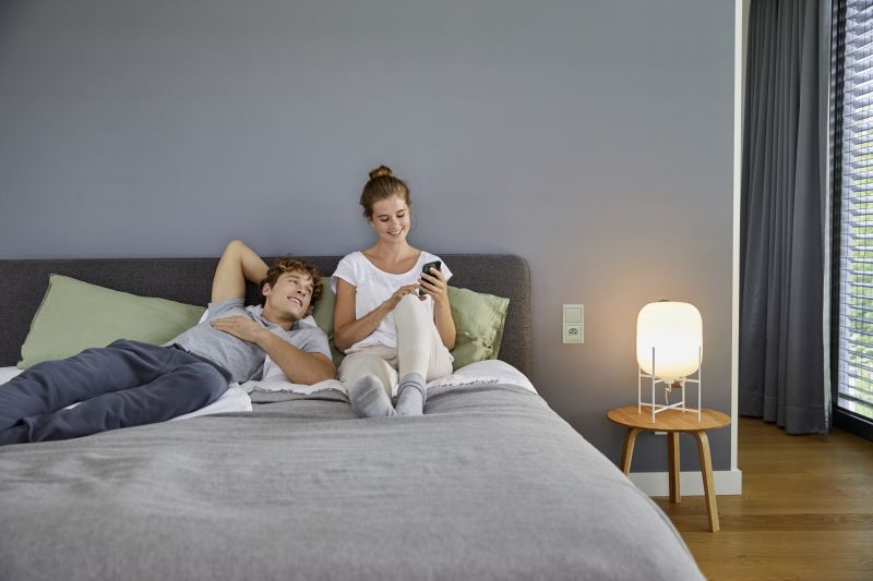 Dwie osoby odpoczywają na łóżku; jedna leży, druga siedzi i korzysta ze smartfona, obok świeci lampka.