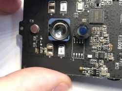 Zbliżenie na czarną płytkę PCB z obiektywem kamery i elementami SMD, trzymaną w palcach