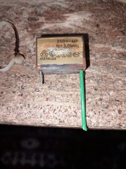 Uszkodzony kondensator 250V~ MP 3,0 MHz leży na drewnianym blacie.
