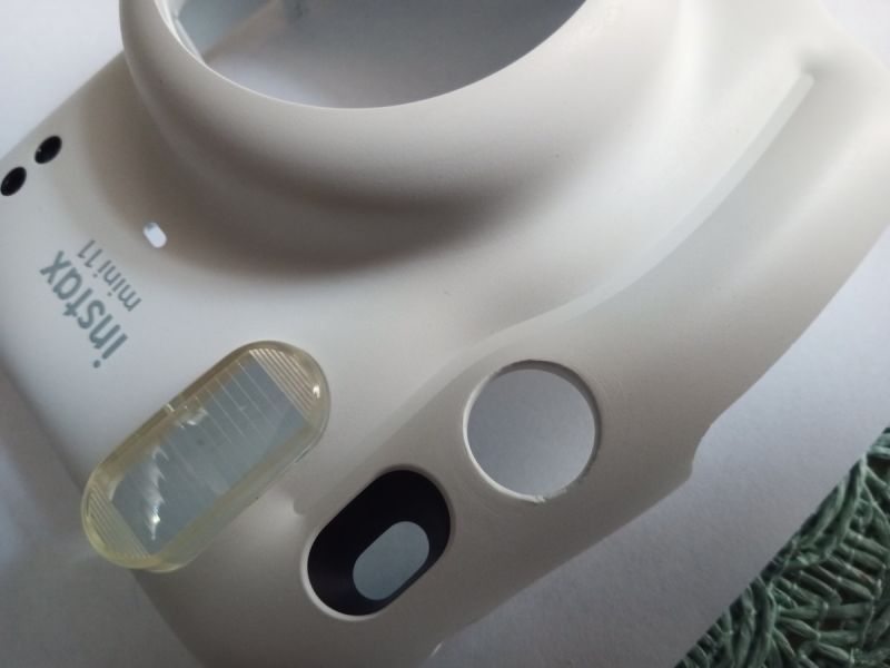 Zbliżenie białego aparatu Instax Mini 11 z obiektywem, lampą i przyciskiem migawki
