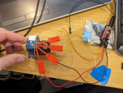 A hand holding an electronic setup with cables and test hooks connected to a USB to TTL converter on a wooden desk next to a laptop.