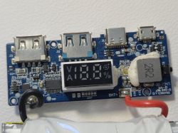 Płytka elektroniczna z wyświetlaczem procentowym oraz kilkoma portami USB, leżąca na białej powierzchni.