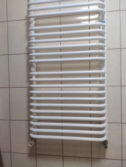 Biały grzejnik łazienkowy na tle jasnych płytek ceramicznych.
