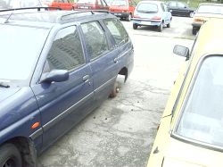 Niebieskie kombi zaparkowane bez tylnego lewego koła na parkingu, obok stoi żółty samochód.