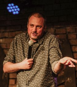 Mężczyzna trzymający mikrofon, ubrany w koszulę w kropki, wykonuje stand-up na tle ściany z cegieł.