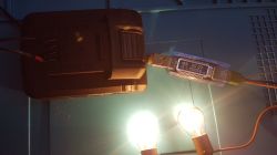 Tester USB z wyświetlaczem podłączony do czarnego akumulatora; poniżej świecą dwie żarówki
