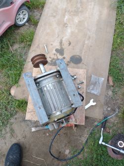 Silnik elektryczny zamocowany na metalowej ramie, ustawiony na drewnianej desce na ziemi, w otoczeniu narzędzi.