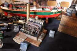 Zestaw modeli silników parowych i modelu statku na stole wystawowym.