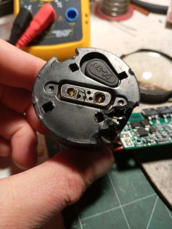 Uszkodzona plastikowa zatyczka akumulatora z gniazdem na styki DC12V, trzymana w dłoni na tle multimetru i narzędzi.