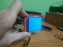 Hand holding a small LCD module with a blue screen on a desk with electronics in the background