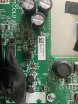 Zbliżenie na fragment zielonej płyty głównej telewizora z widocznymi kondensatorami, rezystorami i naklejką z kodem QR oraz informacjami technicznymi.