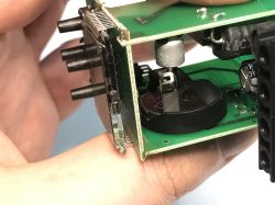 Zbliżenie na płytkę elektroniki z widoczną baterią pastylkową, kondensatorami i złączami.