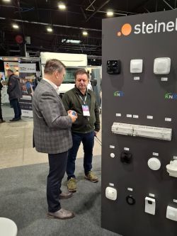 Two men talk at a trade show beside a Steinel display panel with KNX-labeled electrical devices.
