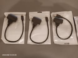 Trzy adaptery do dysków SATA na USB w oryginalnych opakowaniach leżące na szarej powierzchni.