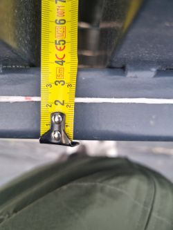 Zbliżenie na stalową belkę bramy mierzoną żółtą miarką, pokazującą odchylenie około 2,5 cm.