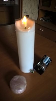 Biała świeca LED z płomieniem, podłączona do baterii R20 i przetwornicy, obok korek silikonowy