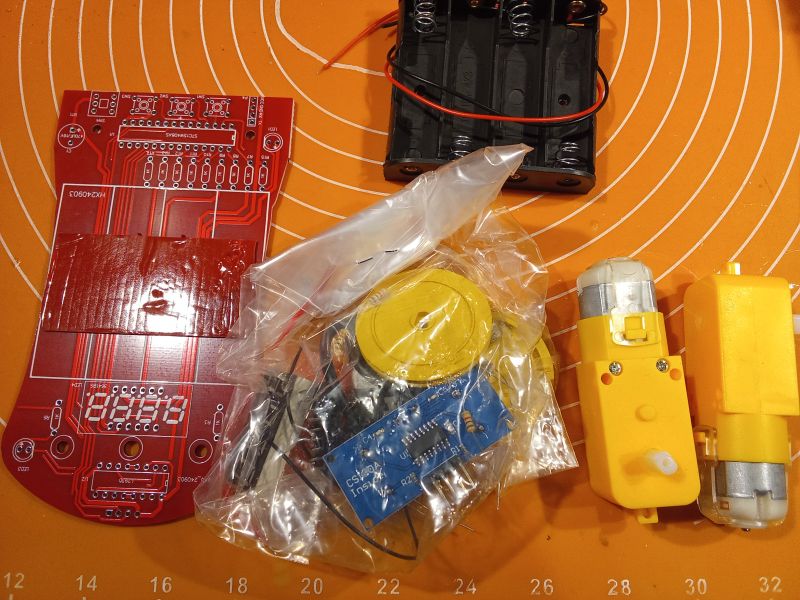 Elektroniczne komponenty na pomarańczowej macie. Widać płytkę drukowaną, koszyk na baterie, dwa żółte silniki i plastikowy woreczek z drobnymi częściami.