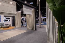 Trade show booth with curtains, roman blinds, and a living-room display; visitors in the background