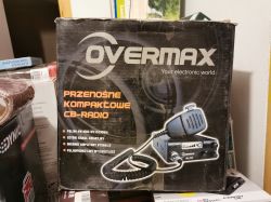 Pudełko z przenośnym kompaktowym CB-radio marki Overmax wśród innego sprzętu elektronicznego.