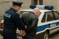 Police officer handcuffing a man in a black jacket in front of FSO Polonez patrol car
