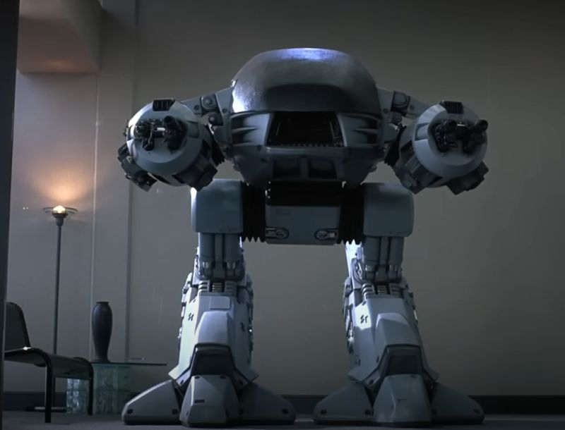 Duży szary robot bojowy z dwoma działkami w ciemnym pomieszczeniu biurowym