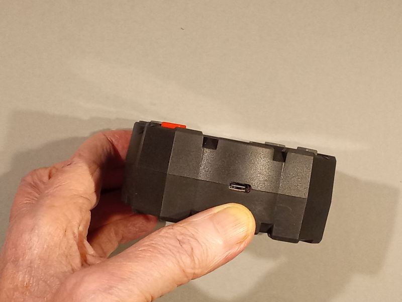 Dłoń trzyma czarne urządzenie z portem micro‑USB na boku, na jasnym tle.