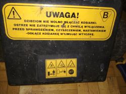 Zbliżenie na żółtą naklejkę ostrzegawczą z instrukcjami bezpieczeństwa na obudowie kosiarki elektrycznej.