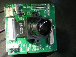 Camera module on a circuit board with a large lens and technical markings.