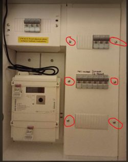 Zdjęcie szafy elektrycznej z licznikiem i płytek z wyłącznikami zabezpieczającymi, z zaznaczonymi śrubami do odkręcenia.