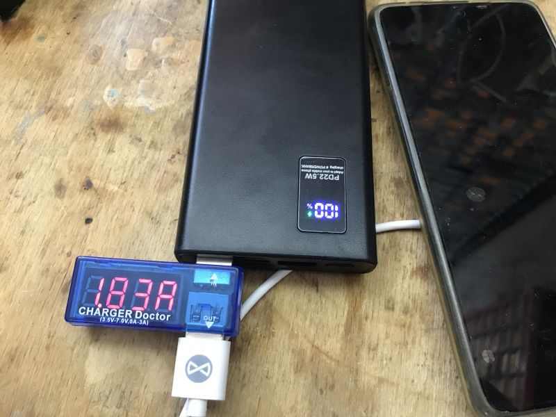 Power bank showing 100% charge, connected to a current meter and a smartphone