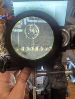 View through an optical eyepiece showing a magnified circuit board area with traces and solder points