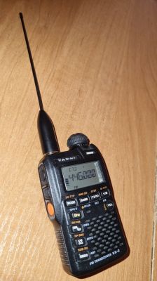 Ręczny transceiver Yaesu VX-3 na drewnianym blacie, z wysuniętą anteną i włączonym wyświetlaczem.