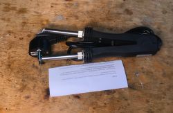 Electric tweezers with a cable and plug lying on a wooden table, next to a sheet of paper.