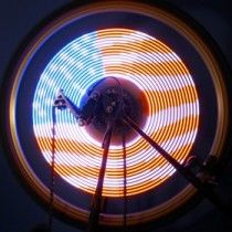 Colorful LED spirals around a bicycle wheel, shot in darkness with visible wires