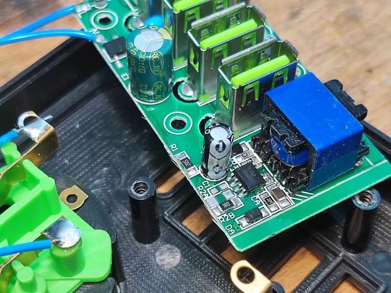 Zbliżenie na płytkę drukowaną wewnątrz listwy zasilającej z pięcioma portami USB, widoczne kondensatory, złącza i elementy elektroniczne.