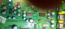Zbliżenie na zieloną płytkę PCB z układami scalonymi, kondensatorami i ścieżkami; u góry częściowo zasłonięta.