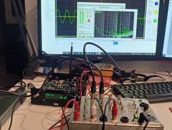 Urządzenie do pomiaru wzmacniaczy audio z podłączonymi kablami, na tle monitora z wykresami.