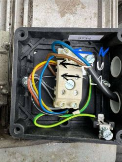 Wnętrze skrzynki elektrycznej z widocznymi przewodami i blokiem zaciskowym; przewody oznaczone strzałkami i kolorami.