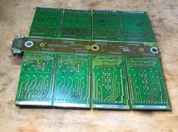 Underside of green PCB assembly with eight modules and a side metal connector on a wooden surface
