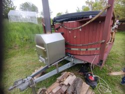 Metalowy piec zewnętrzny przyczepiony do balii ogrodowej z drewnianą obudową na trawniku.